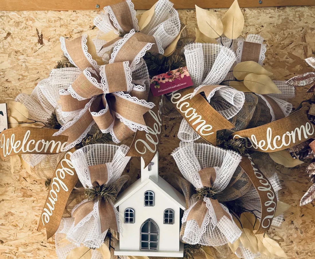 A burlap-and-lace wreath on the workshop wall, wrapped in Welcome ribbons around a little white church.