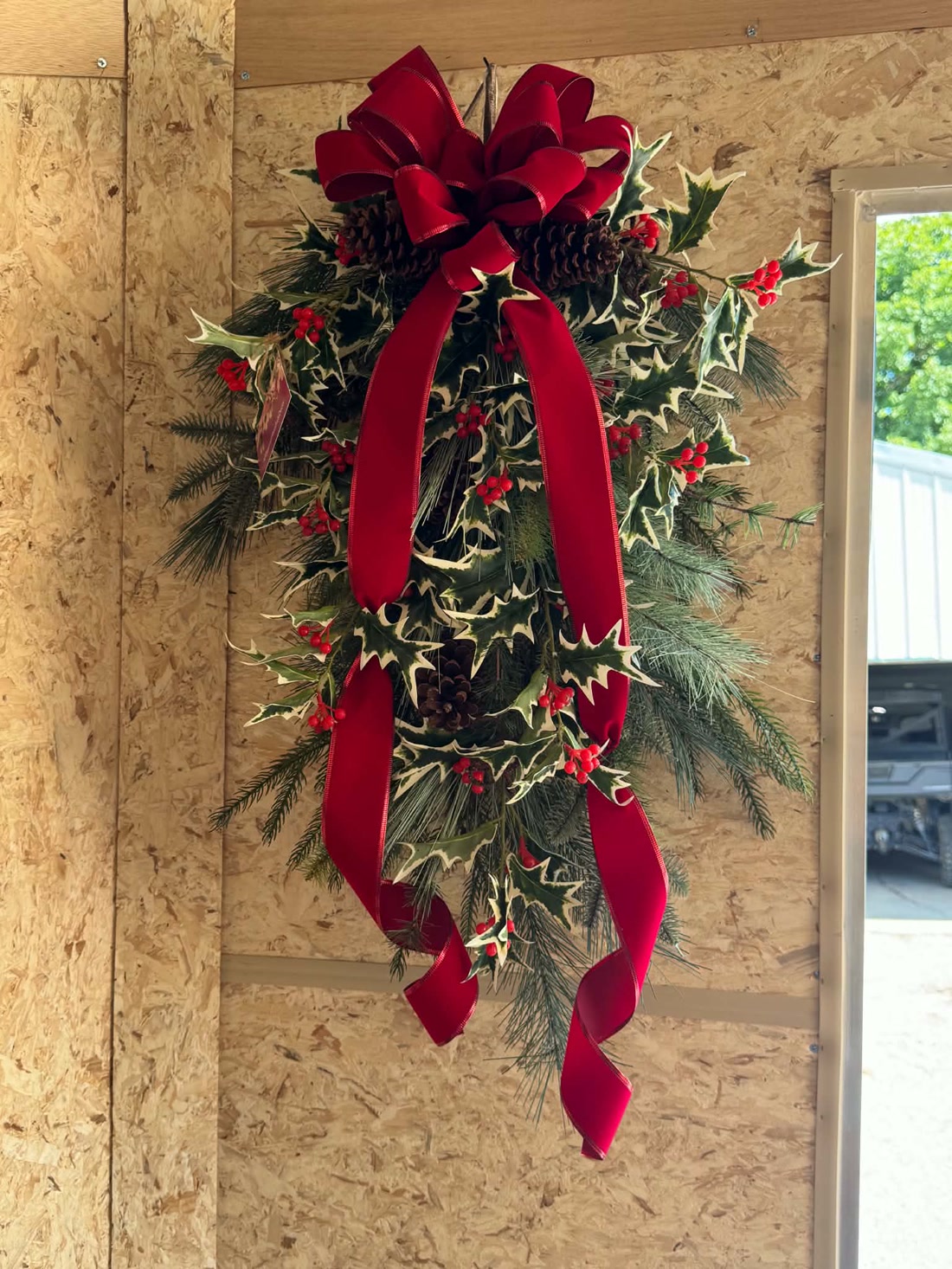 A holly door swag with a deep red velvet bow, pinecones, and trailing ribbon, hanging on the workshop wall.