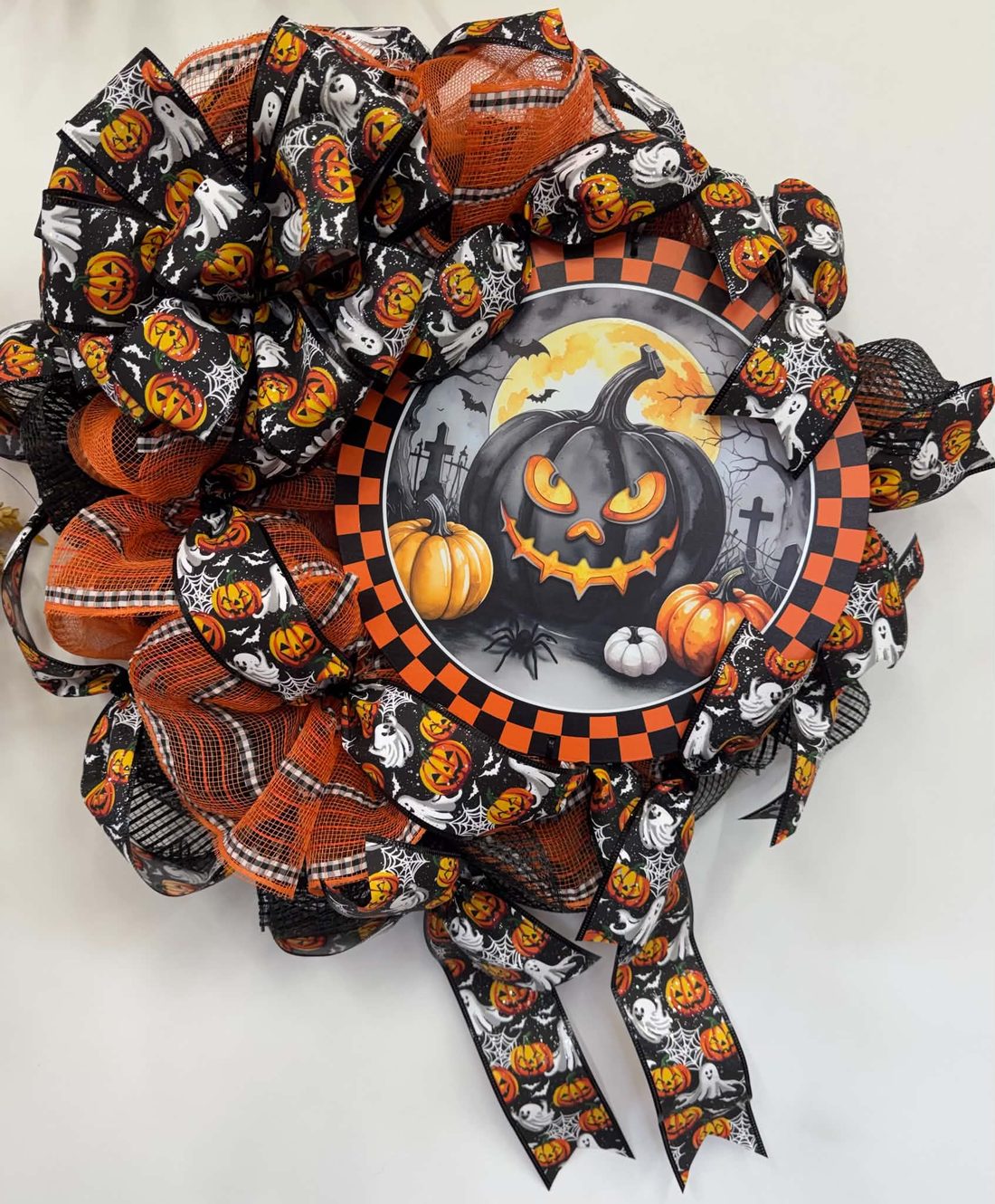 A checker-rimmed sign with a grinning black jack-o'-lantern under a full moon, set in orange mesh and pumpkin-and-ghost ribbon.