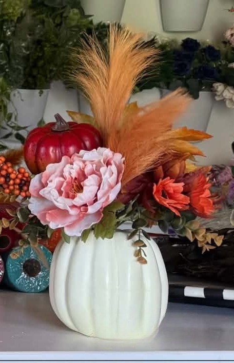 A cream pumpkin vase with a blush peony, coral blooms, a deep-red pumpkin, and tall orange pampas plumes.