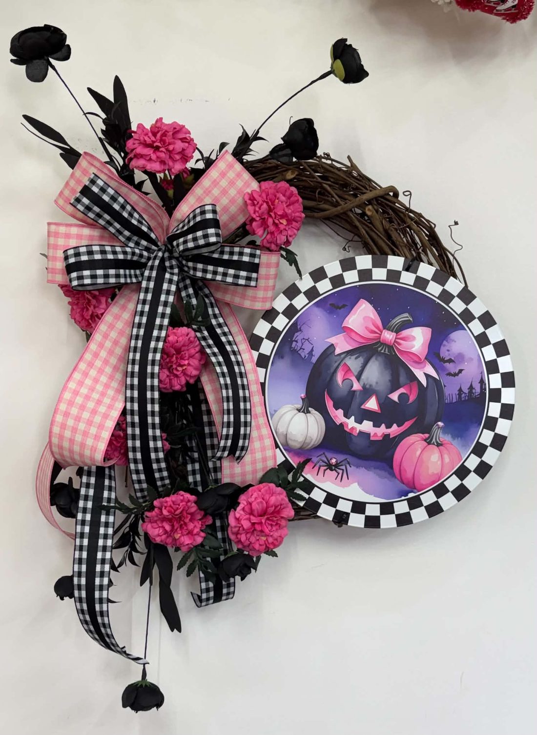 A pink-Halloween grapevine wreath: pink gingham and buffalo-check bow, hot-pink carnations, black roses, and a checker-rimmed pink jack-o'-lantern sign.