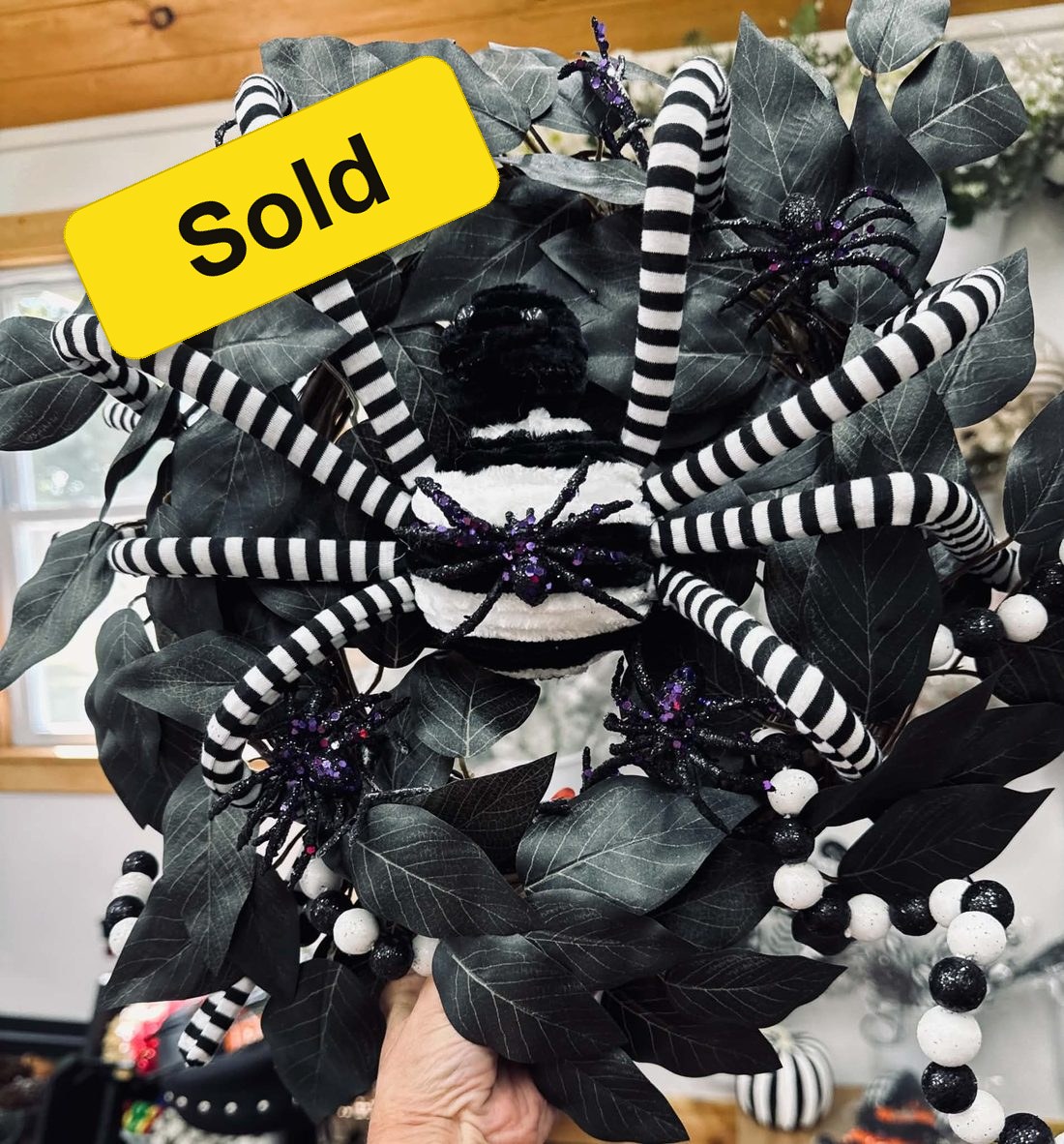 The striped-leg spider wreath with a yellow Sold sticker: a big plush spider with black-and-white striped legs across dark leaves and glitter spiders.