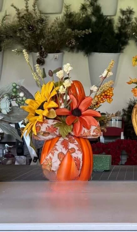 An orange pumpkin vase with yellow sunflowers, rust daisies, cream blooms, and a fall-leaf ribbon bow.