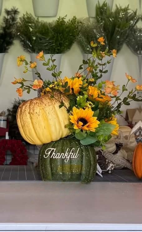 A speckled green 'Thankful' pumpkin vase topped with a big cream pumpkin, golden sunflowers, and sprays of orange blossoms.