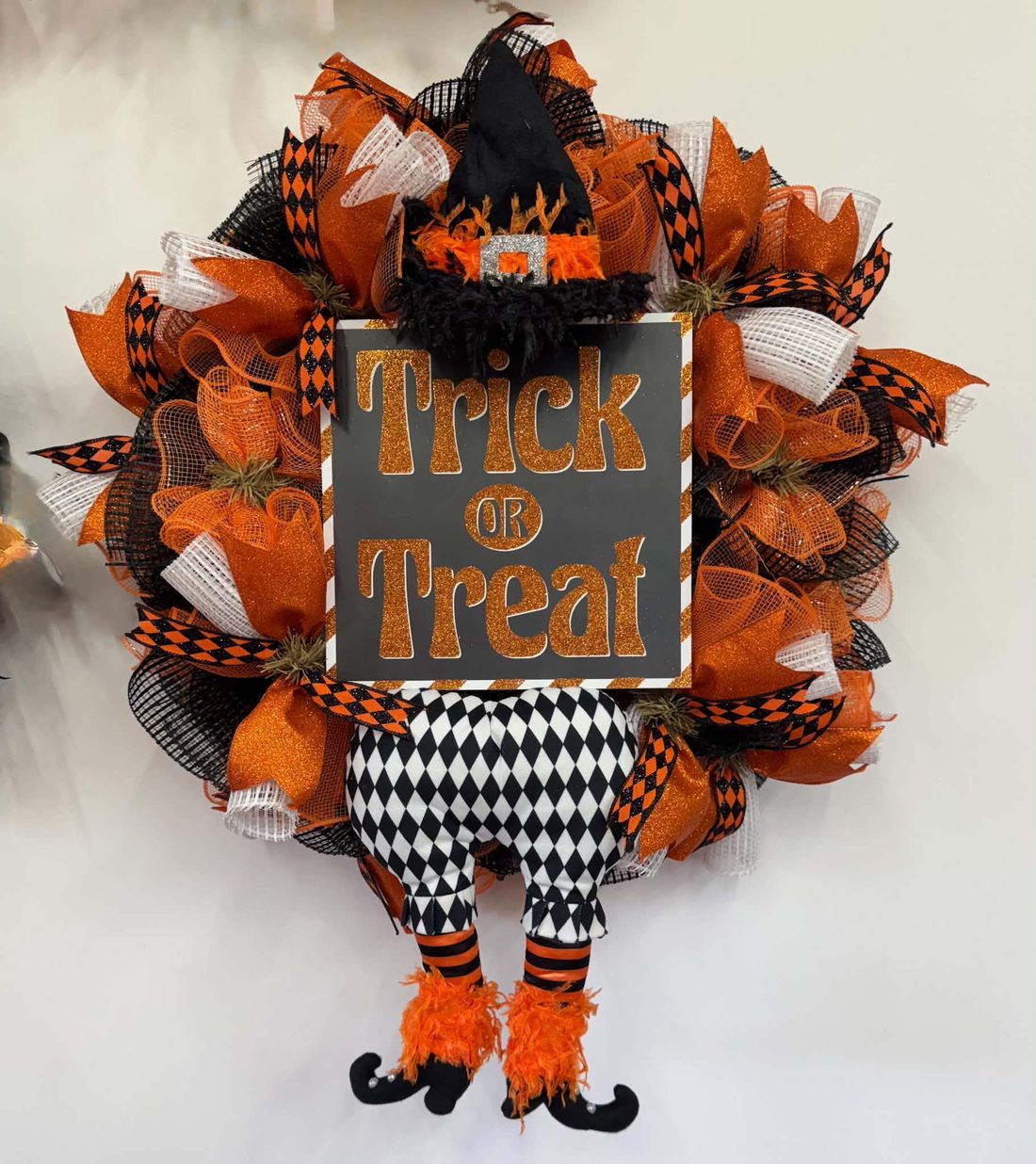 A glittered 'Trick or Treat' sign wearing a witch hat, with harlequin britches and fuzzy orange boots dangling below orange, black, and white mesh.