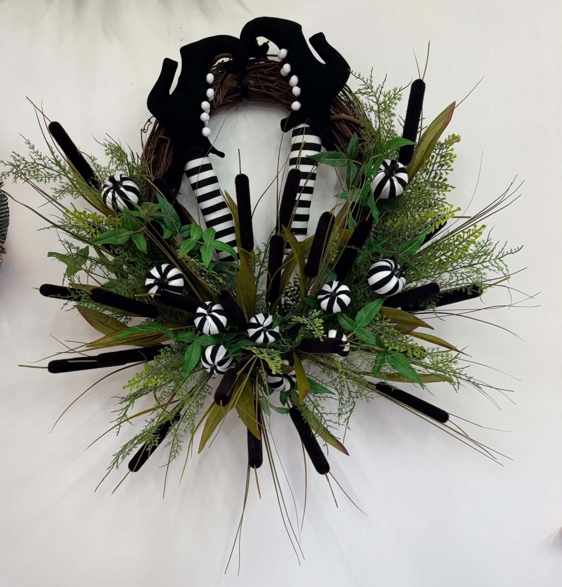 A witch gone in headfirst: striped stockings and curled black boots kicking out of a grapevine wreath full of cattails, wild greenery, and striped mini pumpkins.